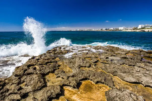 Rocky shore, clear turquoise sea water and the waves break with splashes on the rocks, blue sky. Ayia-Napa, Cyprus.