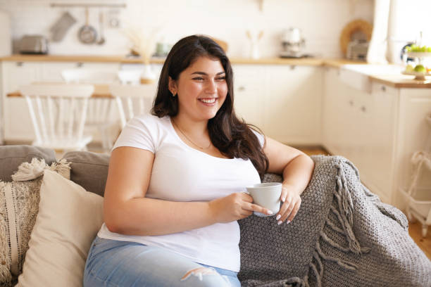 retrato de alegre carismática joven con sobrepeso hembra con pecho grande y pelo negro largo posando en el interior de la cocina elegante, tomando café en el cómodo sofá y riendo de broma divertida - fat fotografías e imágenes de stock