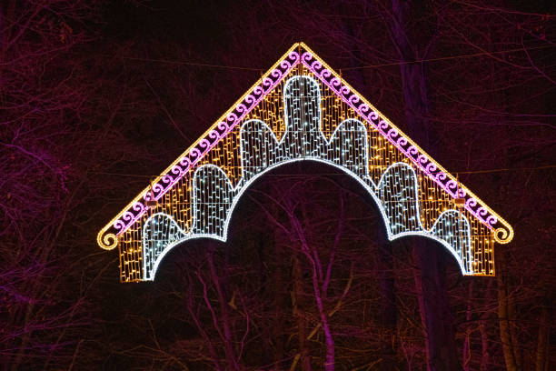 istock Ornate illuminated decoration is hanging in darkness with blurred background of tree branches. Colorful Christmas light decoration in shape of arch. New Year celebration backdrop with copy space. 1190120731