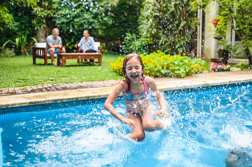 Close-up of laughing young girl with eyes shut jumping into backyard swimming pool and making big splash.