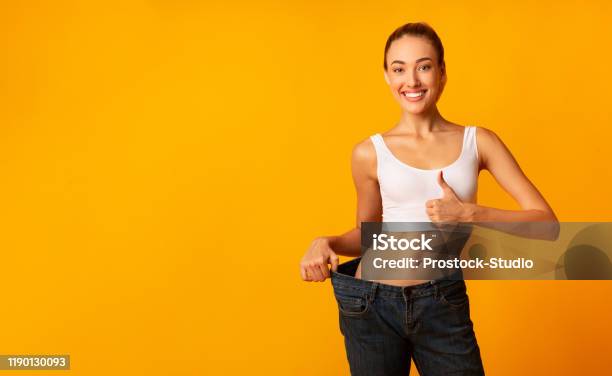 Ragazza In Jeans Oversize Gesticolando Pollici In Su Sorridente Sfondo Giallo - Fotografie stock e altre immagini di Dieta