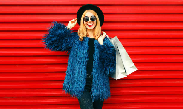 青いフェイクファーコート、黒い丸い帽子、赤い壁の背景にポーズをとるサングラスを身に着けたショッピングバッグを持つ肖像画スタイリッシュな笑顔の女性 - coat ストックフォトと画像