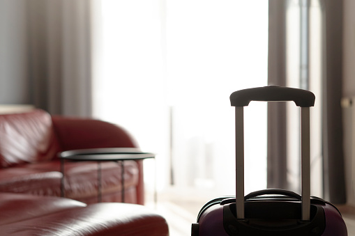 A suitcase standing by the window in the living room near a large leather red sofa and an empty table. The concept of moving and starting a new life in a new apartment. No people interior