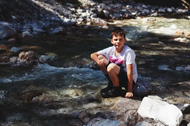 Lofer, Austria - August 11, 2019: teenager boy sitting happy on a rock near cold mountain stream in Austrian Alps and having rest after a difficult and hot hike up the mountain
