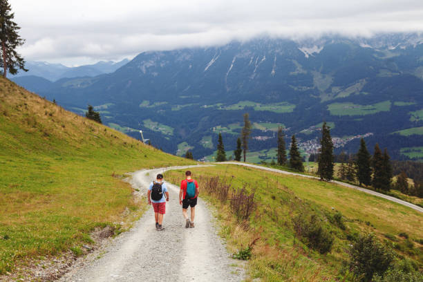 Rear view of a father and teenage son walking on a footpath high up in Alps, Austria, while on their hiking trip with beautiful panoramic views and mountains in distance. Lofer, Austria - August 10, 2019: a man and his teenager boy walking a dirt road high up on a mountains ridge in Alps, Austria, while carrying their backpaks and having a conversation in an amazing setting with more mountains in the horizon single lane road footpath dirt road panoramic stock pictures, royalty-free photos & images