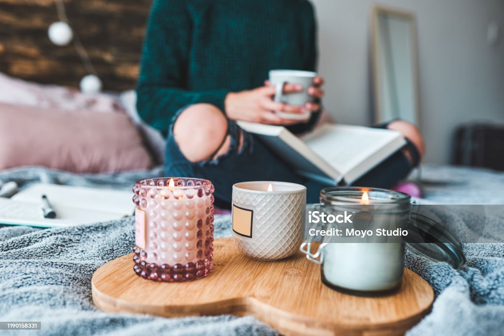 Creating a relaxing environment like no other Cropped shot of an unrecognizable young woman relaxing with a book and a cup of coffee on her bed at home Candle Stock Photo