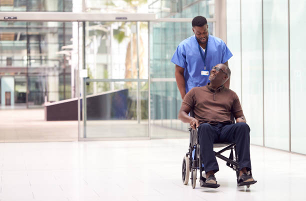 male nurse wearing scrubs wheeling patient in wheelchair through lobby of modern hospital building - male nurse black nurse doctor imagens e fotografias de stock