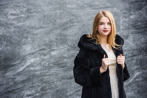 Fashion portrait of young blonde elegant woman in black dress and black fur coat at city street