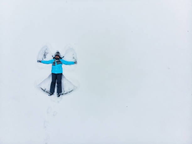 Close up front high angle shot of young smiling Asian adult woman, wearing warm winter clothing, laying down or lying down in thick heavy snow and playing with it, feeling cold, while exploring the nature in mountains.Young happy Asian female traveler or tourist having fun and relaxing on a solo-trip or travel in Japan during winter holidays, enjoying her day outdoors.
