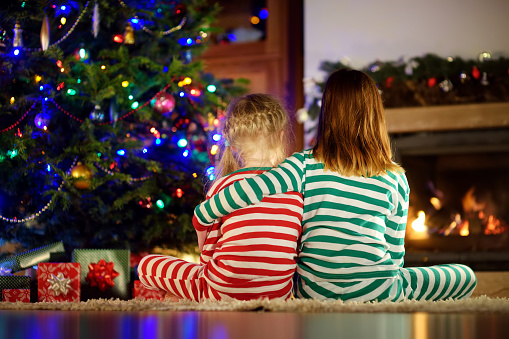 Hermanas pequeñas felices que llevan pijamas de Navidad abriendo cajas de regalo junto a una chimenea en una acogedora sala de estar oscura en la víspera de Navidad. photo