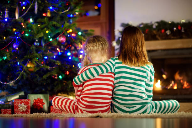 hermanas pequeñas felices que llevan pijamas de navidad abriendo cajas de regalo junto a una chimenea en una acogedora sala de estar oscura en la víspera de navidad. - warming up fotos fotografías e imágenes de stock