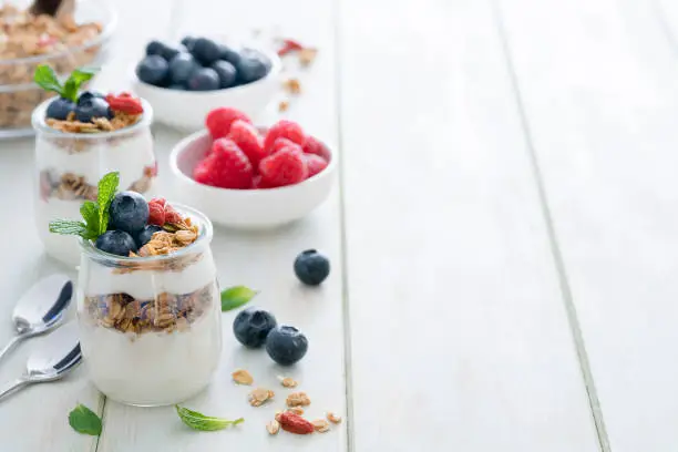 Healthy eating: two glass containers with homemade yogurt and granola shot on breakfast table. Two spoons are beside the yogurt containers. Some berries complete the composition. The composition is at the left of an horizontal frame leaving useful copy space for text and/or logo at the right. Predominant color is green. High resolution 42Mp studio digital capture taken with Sony A7rii and Sony FE 90mm f2.8 macro G OSS lens