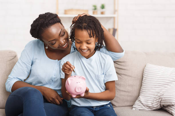 Little black girl putting money to piggy bank African Mother And Daughter Putting Coins Into Piggy Bank At Home. Free space financial education stock pictures, royalty-free photos & images