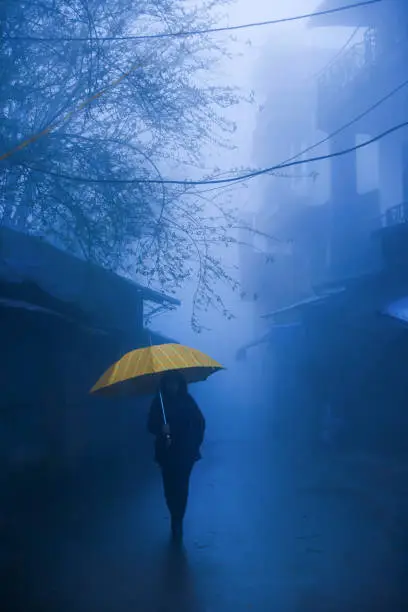 An unidentified female walks with umbrella in the dark blue foggy. Lonely woman in black jacket walks with yellow umbrella on the alley in an old town.