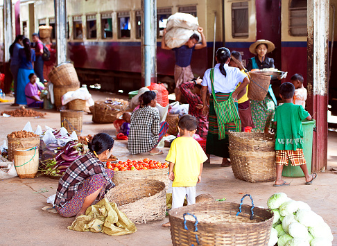Sung Ban railway station platform, near Kalaw, Shan State, Myanmar. A  group of agriculture farm workers and families dressed in shabby working clothes and hats sitting huddled together or working companionably on the platform of Sung Ban railway station, with their produce displayed for the wholesale vegetable market, with train carriages and freight cars behind.