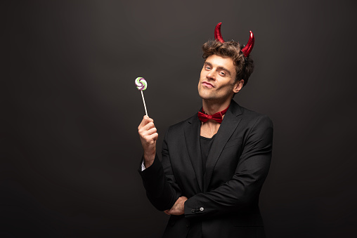 handsome man in halloween devil costume holding one lollipop on black