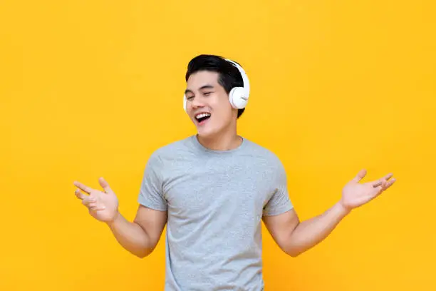 Happy young Asian man wearing headphones listening to relaxing music with hands open isolated on yellow background