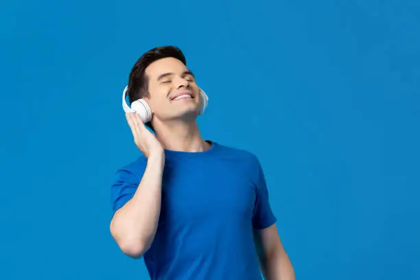 Relaxing young man listening to music from headphones with eyes closed isolated on blue background