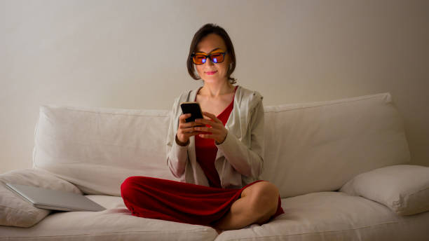 Young woman is sitting on white sofa looking at the smartphone with yellow (amber) blue light blocking eye glasses - health and wellness concept for melatonin and circadian rhythm stock photo