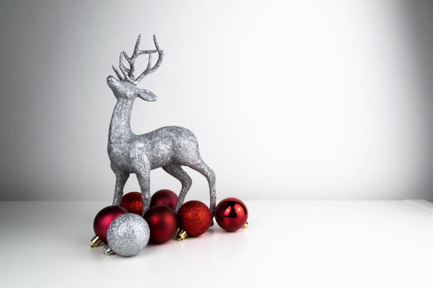 istock Silver glitter coated Reindeer Table Top Christmas ornament stood up on a white reflective surface and surrounded by shiny red and silver Christmas tree baubles. 1190209645