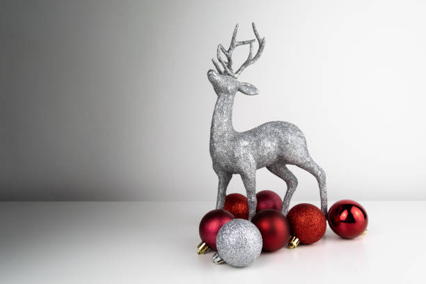 istock Glitter coated Reindeer Table Top Christmas ornament stood up on a white surface, surrounded by shiny red and silver Christmas tree baubles, situated to the right of the image. 1190209772