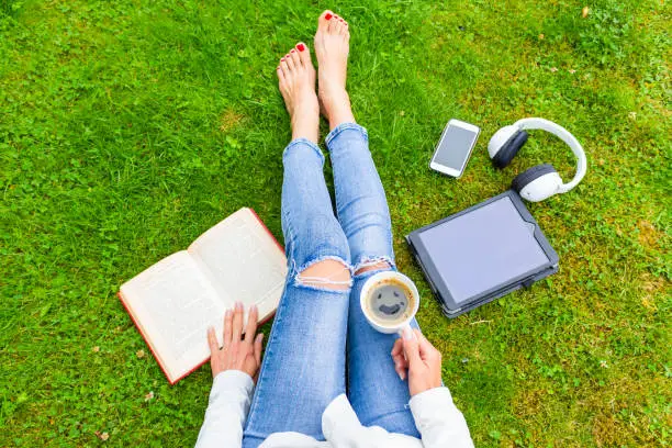 Top view of woman sitting on the green grass, relaxing with smartphone, headphones, tablet, book and coffee (with smile) in the hand