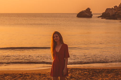 Young smiling blonde girl in red dress walking in the evening on the beach near the sea