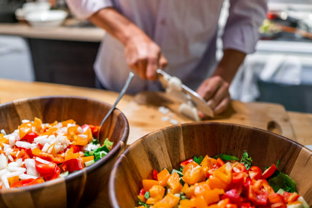 cebolas de sino de corte do homem na tabela de placa de corte no fundo com duas grandes bacias da salada vegan fresca com pimentas de sino alaranjadas vermelhas na cozinha - chopped green bell pepper pepper bell pepper - fotografias e filmes do acervo