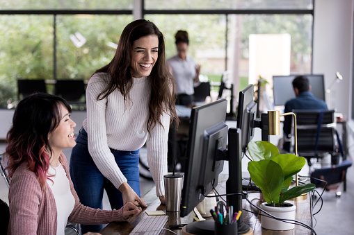 Attractive young businesswoman laughs with a female colleague while working together. They are looking at something on a desktop computer.