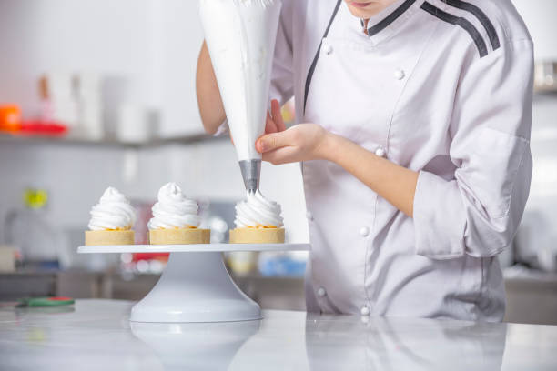 Female chef with confectionery bag squeezing cream on cupcakes.