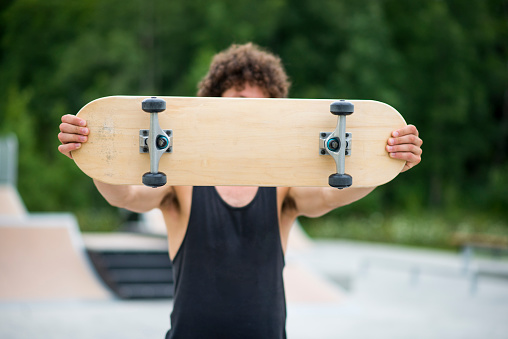 Anonymous young man wearing a black shirt holding up a blank wood skateboard in front of his face while at a skateboard park in the city during summer.