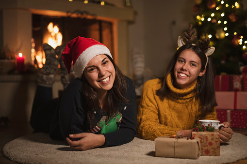 Two happy sisters laying on carpet at home around fireplace and Christmas decoration, wearing sweater and Santa hat, looking at camera