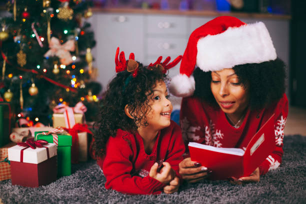 a matriz africano-americana e sua filha pequena que lêem um livro por uma árvore de natal na sala de visitas no inverno - dark little girls child happiness - fotografias e filmes do acervo