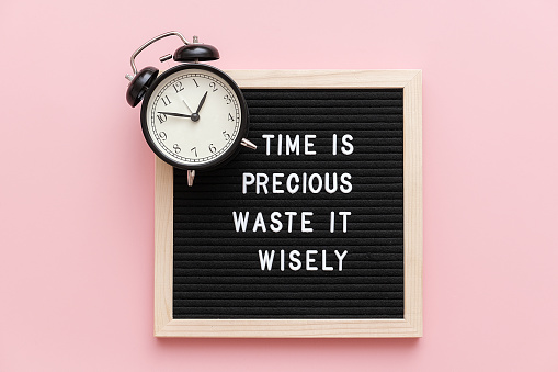 Time is precious waste it wisely. Motivational quote on letterboard and black alarm clock on pink background. Top view Flat lay Concept inspirational quote of the day.