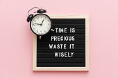 Time is precious waste it wisely. Motivational quote on letterboard and black alarm clock on pink background. Top view Flat lay Concept inspirational quote of the day