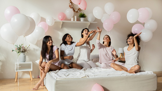 Beautiful happy multiethnic ladies having fun playing with pink balloons in bedroom, excited young girls friends wear pajamas celebrate birthday bachelorette slumber party sitting on bed together