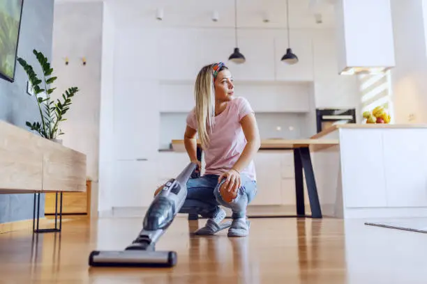 Worthy caucasian blond housewife crouching in living room and using steam cleaner to clean parquet.
