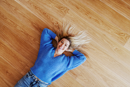 Top view of attractive charming smiling caucasian blond young woman in sweater lying on parquet and looking at camera.