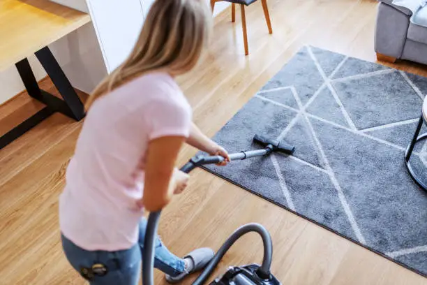 Rear view of attractive worthy caucasian young housewife using vacuum cleaner to clean carpet in living room.