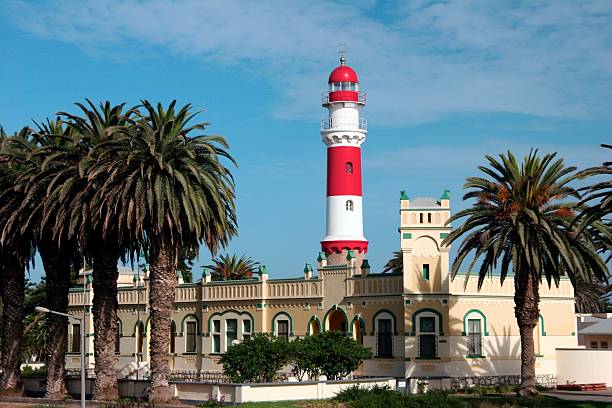 A DSRL photo of a lighthouse in Itapuã Beach, Salvador, Bahia, Brazil. The lighthouse is painted with white and red stripes and has a walkway leading to the road. The day is sunny with scattered white clouds. The sky has a gradient from dark blue to light blue.