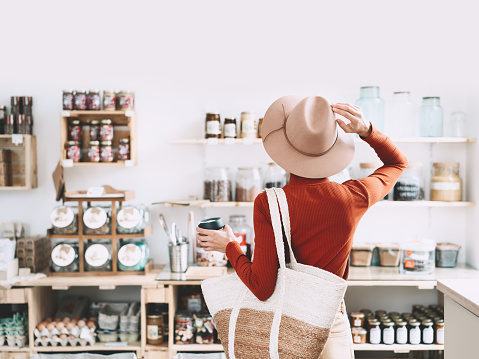 Minimalist vegan style girl with wicker bag and reusable glass coffee cup on background of interior of zero waste shop. Woman doing shopping without plastic packaging in plastic free grocery store.