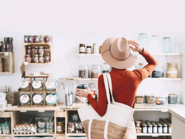 Photo of Minimalist vegan style girl with wicker bag and reusable glass coffee cup on background of interior of zero waste shop.