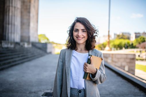 Close-up of relaxed female law student in casual attire standing with book at entrance to university in Buenos Aires.