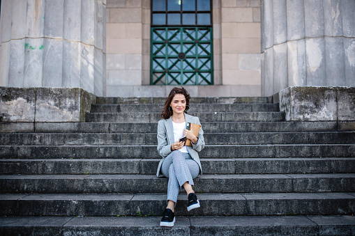 Outdoor portrait of smiling young female law student sitting with book and smart phone on steps to university building.