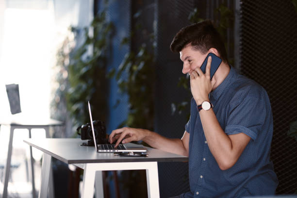 istock Young male freelancer sitting in a cafe and working on the computer, talking with his smartphone indoors with casual dressing. Young businessman spending time on the computer alone. 1190367578