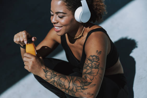 Joyful Afro American woman in headphones holding bottle of juice Smiling young lady listening to music outdoors stock photo black tattoo stock pictures, royalty-free photos & images