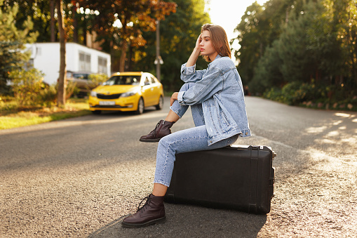 Profile portrait pensive girl sits on her suitcase on middle road, waiting a transportation, in sunset background. Horizontal view.