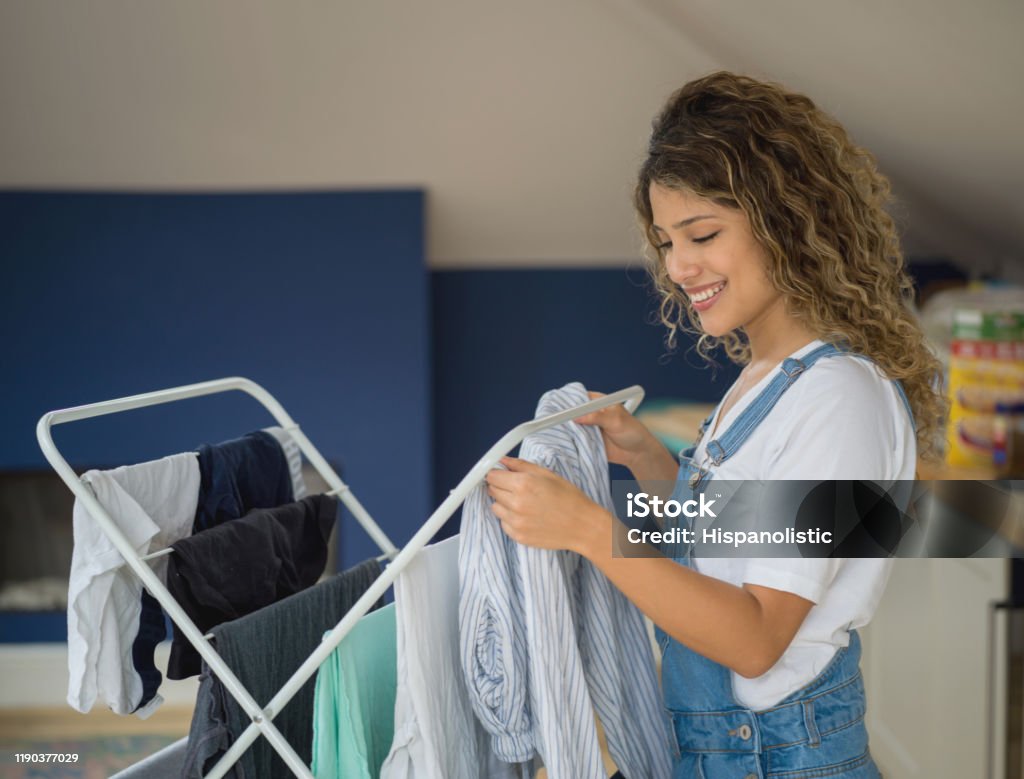 Young woman hanging the laundry on drying rack smiling at home Young woman hanging the laundry on drying rack smiling at home - Lifestyles Clothing Stock Photo