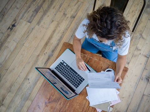 High angle view of young woman at home using her laptop holding bills and credit card - Lifestyles