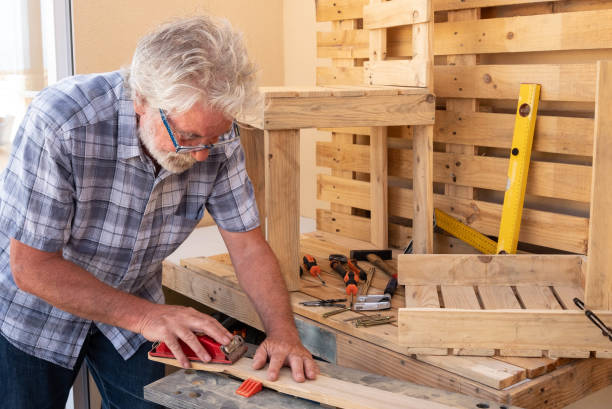 Senior man with white hair enjoying work with wooden pallet to create new objects for home. A chair is almost finished and another is in work. Free time well spent Senior man with white hair enjoying work with wooden pallet to create new objects for home. A chair is almost finished and another is in work. Free time well spent plane hand tool photos stock pictures, royalty-free photos & images